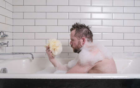 Sad man bathes in a bath with lush soap suds, he holds a sponge in his hands and looks thoughtfully.の写真素材