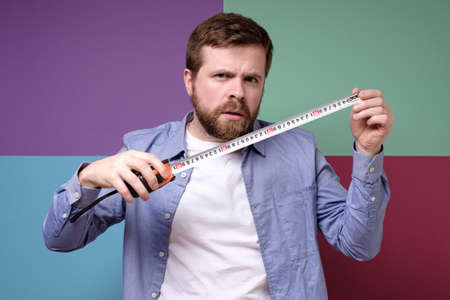 Puzzled, pensive Caucasian man with a construction tape measure in hands. Colorful background. Home repairs.の写真素材