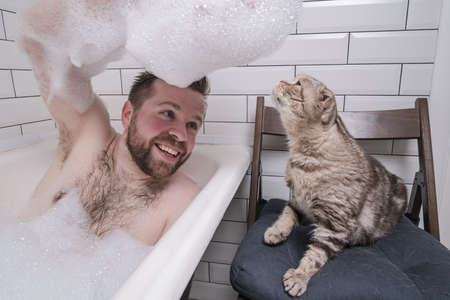 Satisfied man bathes in a bathtub, he holds on hand a lush soap suds, which his adored cat is interested in.の写真素材