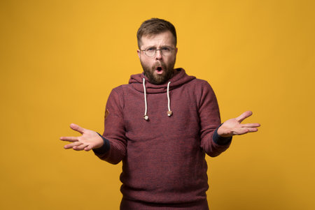 Disgruntled, annoyed bearded man with glasses throws up hands in bewilderment and says something while looking at the camera.の写真素材