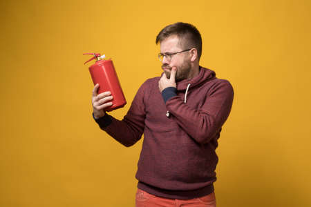 Puzzled man with glasses holds a fire extinguisher in hand and looks at it thoughtfully. Yellow background.の写真素材