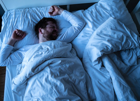 Man in white pajamas has a terrible nightmare, wrinkles face and prepares a fist for protection. Insomnia and mental health concept.の写真素材