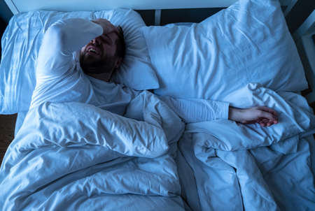 Caucasian bearded man in pajamas suffering from a headache, he put hand on his forehead and closes eyes while lying in bed.の写真素材