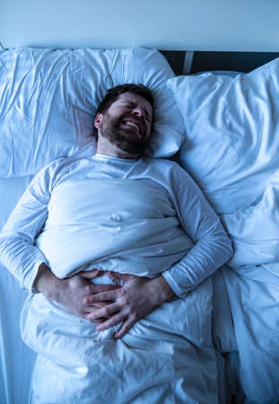 Young bearded man in pajamas suffers from abdominal pain, he holds hands on belly, clenches teeth and closes eyes.の写真素材