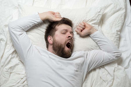 Cute bearded man wakes up from sleep, he yawns and stretches as he lies in bed. Top view. Close-up.の写真素材