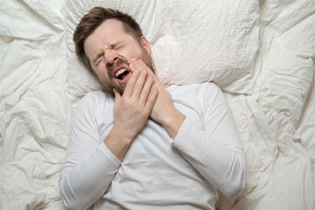 Man suffers from unbearable toothache, he holds hands on cheek and groans in pain, closing eyes and...