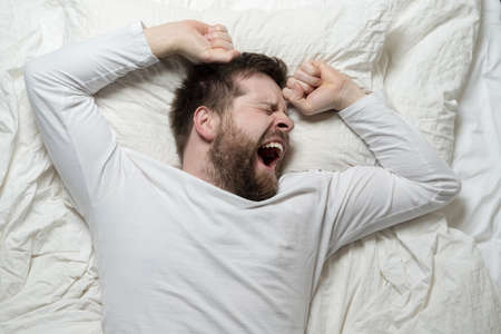 Bearded man in white pajamas is tired and wants to sleep, he yawns and stretches while lying in bed....