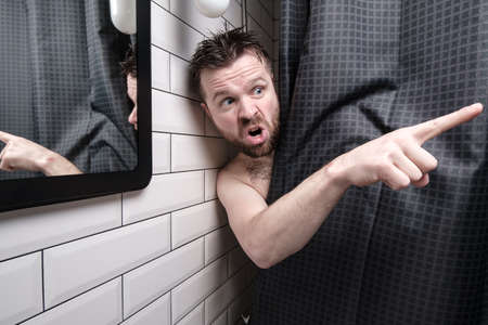 Man is furious, peeking out from behind a shower curtain, he caught someone peeping at him, points a finger at him and yells angrily.の写真素材