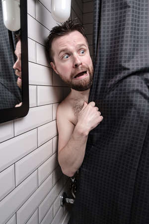 Wet, frightened man peeps out from behind the shower curtain and looks uneasy with a strange expression on his face.の写真素材