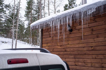 Icicles hang from the roof of a log house near which a car is carelessly parked, which can be damaged by falling ice floes.の写真素材