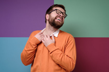 Caucasian bearded man suffers from allergies, he irritably scratches neck, closes eyes and bares teeth. Colorful background.の写真素材