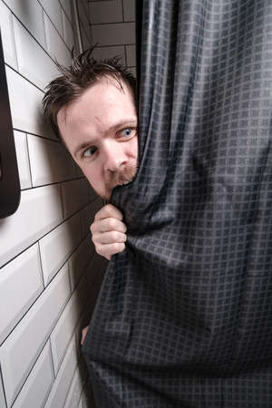 Alarmed, wet man looks out of the shower, bites the curtain with excitement and looks away in dismay.の写真素材