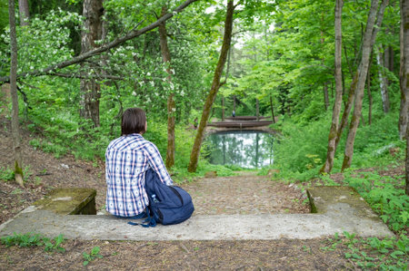 Calm woman with a city backpack sits on the stone steps enjoying the beautiful landscape, pond and forest, in the park.の写真素材