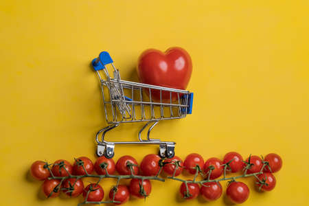 Red tomato in the form of a heart in a grocery trolley, next to cherry tomatoes, on a yellow background. Healthy food concept.の写真素材