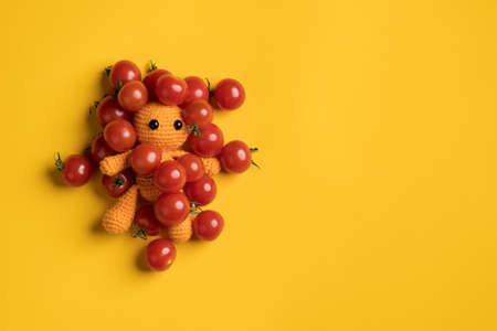 Cute homemade knitted toy man in red cherry tomatoes, on a yellow background. Copy space. Top view.の写真素材
