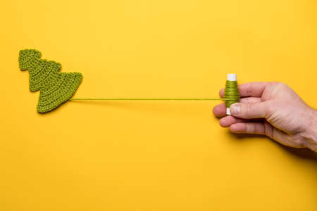 Knitted Christmas tree - a symbol of the New Year and a hand unwinding a spool of thread, on a yellow background. Copy space.の写真素材