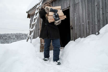 Man carries an armful of firewood against the backdrop of a barn and a snow-covered forest, on a winter day, in the village.の写真素材