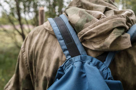 Blue small backpack on a mans back in camouflage clothes, against a blurred background of green bushes.の写真素材