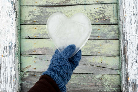 Valentines Day. Heart, a symbol of love, made of ice, is held by a hand in mittens against the background of an old wooden wall.の写真素材