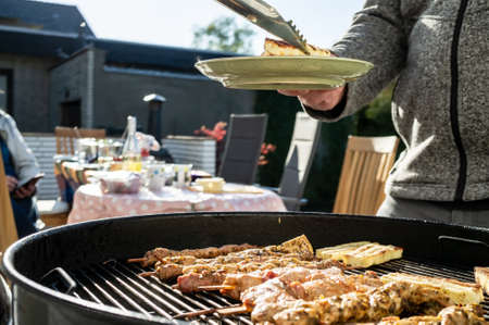 Woman with a plate and tongs for hot food stands near the barbecue with grill meat and cheese, in the courtyard of the house.の写真素材