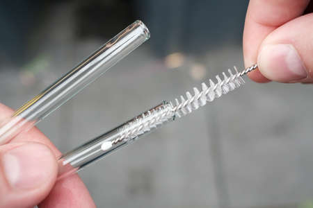 Hands holding reusable glass straws for cocktails and cleaning them with a special brush on a blurred gray background. Ecological concept.の写真素材