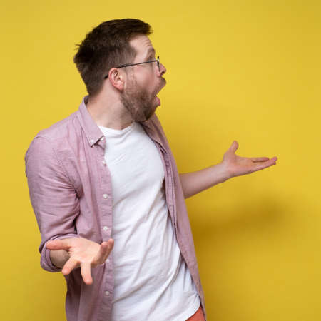 Man is bewilderment and shocked, he looks to the side with his mouth open and spreads his arms in amazement. Yellow background.の写真素材