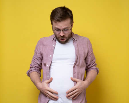 Man is shocked by his overweight, he looks at the excess fat on stomach and holds it with hands. Healthy lifestyle concept.の写真素材
