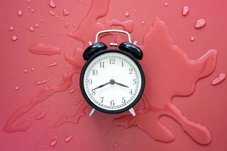 Black retro alarm clock in a pool of spilled water on a red background. Top view.の写真素材