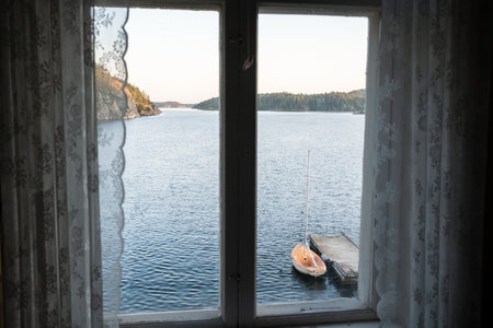 Old closed window across with great views of the sea, rocky shore, moored boat and sky at sunset.