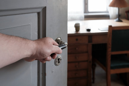 Hhand holds on to the handle on an open wooden door, through which one can see an office, an antique table, a chair, a lamp and a window.の写真素材