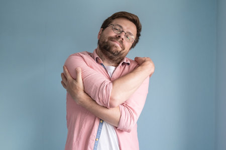 Dreaming about love, a man in glasses and a shirt gently hugs himself, standing on a blue background.の写真素材