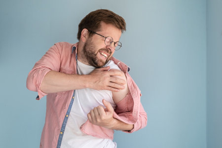 Man is experiencing severe pain in shoulder, he massages the sore spot, closes his eyes and bares his teeth in suffering. Medical concept.の写真素材