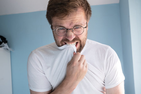 Man with glasses is furious, he tries to contain anger and bites his t-shirt. Concept of psychological health. Close-up.の写真素材