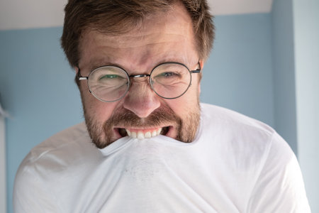 Man with glasses is angry, he tries to contain rage and bites his t-shirt. Concept of mental health. Close-up.の写真素材