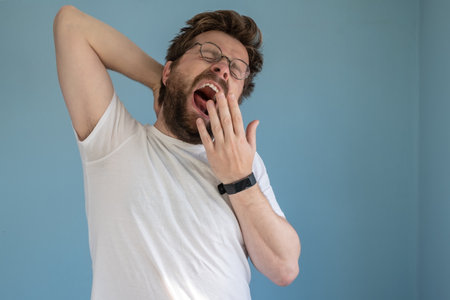 Bearded man in glasses and a white t-shirt yawns with mouth wide open, covering it with hand, he is sleepy or bored. Blue background.の写真素材