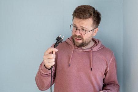 Man with bewilderment looks at a faulty electric plug with a cord. Blue background.の写真素材