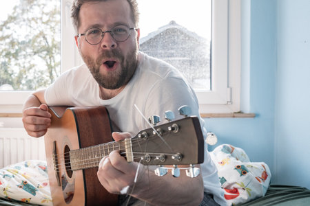 Man plays an acoustic guitar and sings loudly, sitting on a bed.の写真素材