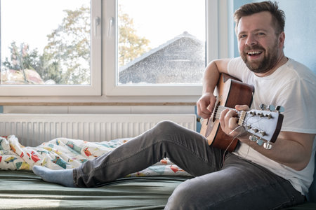 Man plays the acoustic guitar and sings while sitting on the bed.