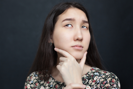 Beautiful Asian girl looks up thoughtfully, doubting the decision. She rubs her chin in uncertaintyの写真素材