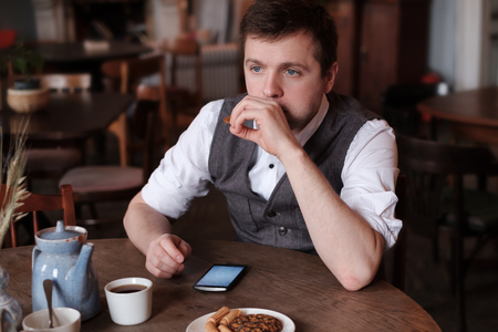 A young man in a white shirt and waistcoat breakfasts a cup of tea with a biscuit and thinks about the plans for the dayの写真素材