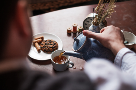 The man pours a cup of hot tea. Sweets lie on the table. He cooks his own breakfastの写真素材