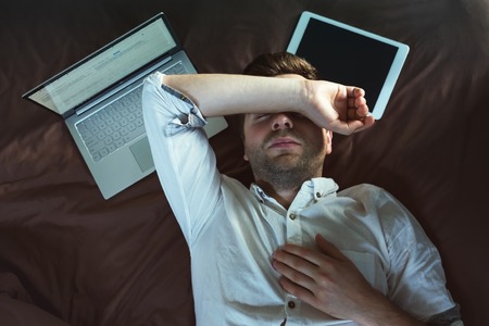 Tired caucasian young man in white shirt sleeping and keeping one hand above head, lying on bed near...