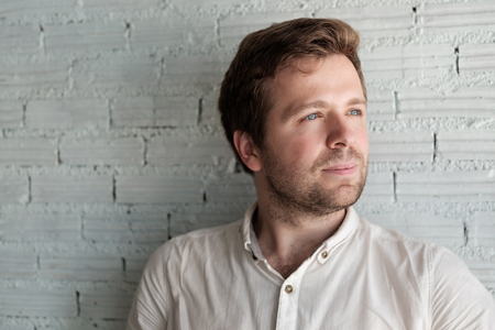 Caucasian man in white shirt standing near wall and smile a little. He is content and confident.