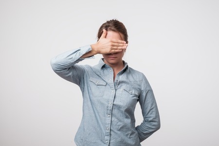 Young european woman in shirt hides her face, studio photo isolated on a gray background. She is sad and ashamedの写真素材