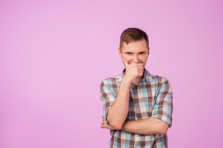 Shot of attractive young caucasian man looking at camera with cunning funny expression, closing his mouth. He is trying not to laugh standing on pink backgroundの写真素材