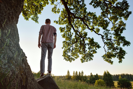 A man in checkered shirt standing near tree and looking aside on valley.の写真素材