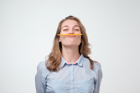 Young beautiful student holding pen between nose and lips as mustache looking funny and naughty after studying long hours isolated on white backgroundの写真素材
