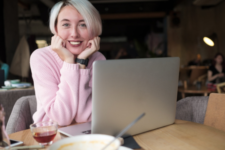 Happy smiling young female student using laptop at library or coffee shop. Cheerful hipster girl working on portable computer at vintage loft.の写真素材