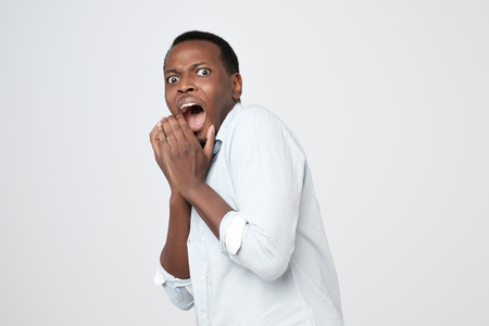 Puzzled african man isolated on grey background dressed in white shirt, covering mouth with handsの写真素材