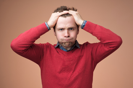 Upset unhappy young man in red sweater squeezing head with hands, suffering from headache. People, stress, tension and migraine conceptの写真素材
